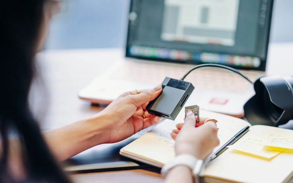 Eine Person steckt eine SD-Karte in ein Kartenlesegerät. Sie sitzt am Schreibtisch, im Hintergrund sind ein Bildschirm und eine digitale Spiegelreflex-Kamera zu sehen.